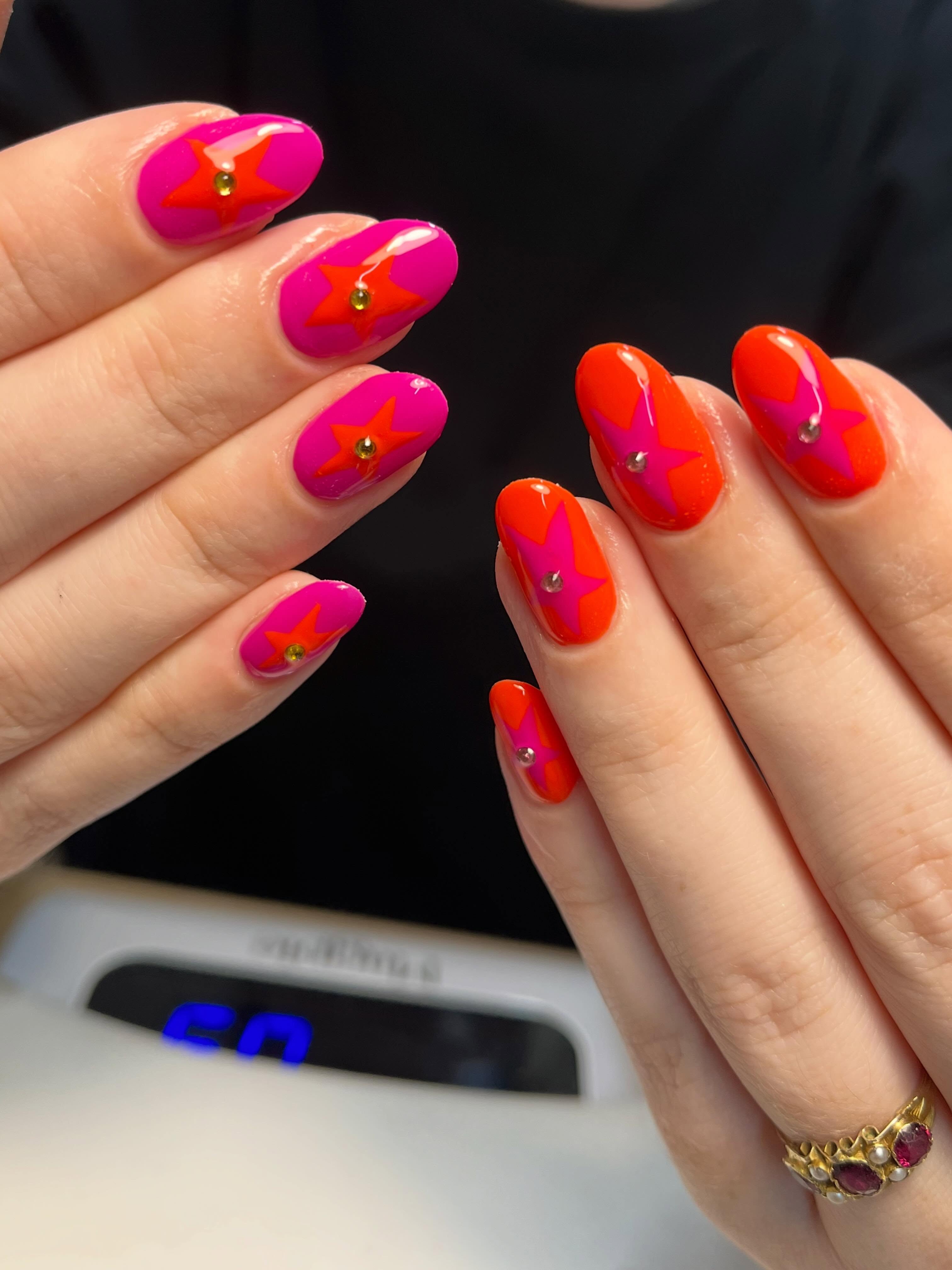 Neon pink and orange contrasting star nails with a glossy finish and crystal embellishments on natural oval-shaped nails
