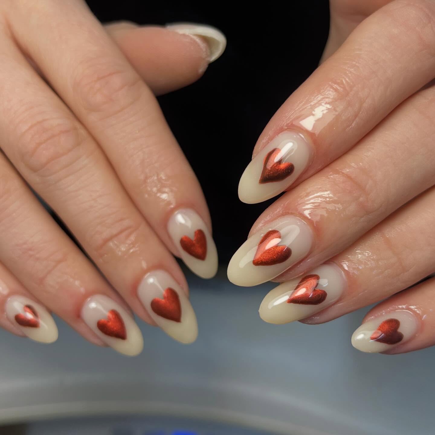 Cream almond nails with metallic red chrome heart accents, an elegant and romantic statement manicure